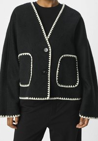 Svart fleece cardigan med vit vågig kant, med stora framfickor och avslappnad silhuett. Stängs med två svarta knappar.