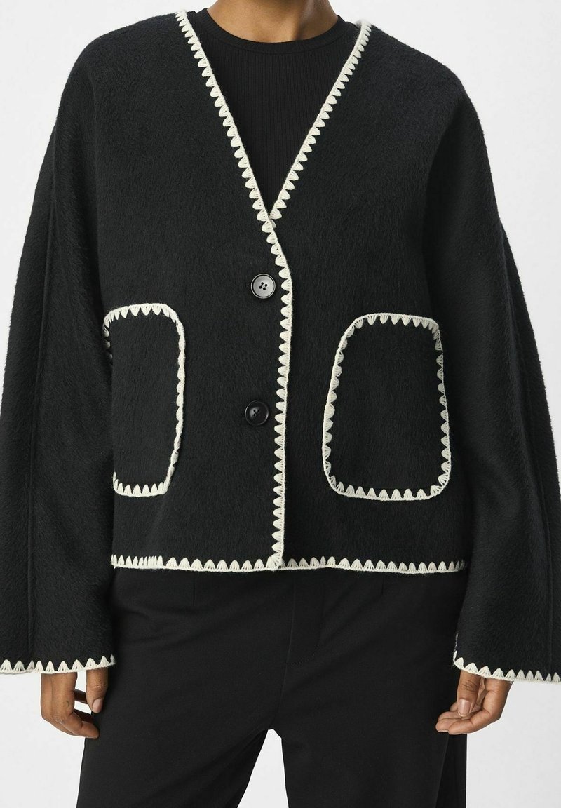 Svart fleece cardigan med vit vågig kant, med stora framfickor och avslappnad silhuett. Stängs med två svarta knappar.