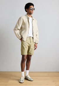 Ελαφρύ μπεζ μπουφάν bomber πάνω από λευκό μπλουζάκι, σε συνδυασμό με σορτς σε ανοιχτό ελιάς και αθλητικά παπούτσια με πράσινες λεπτομέρειες. Φοράει σκουρόχρωμα γυαλιά ηλίου.