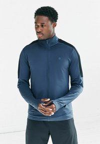 Granatowa sportowa kurtka na zamek błyskawiczny z czarnymi akcentami na ramionach, długimi rękawami z otworami na kciuki i dopasowanym krojem.