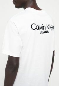 White cotton t-shirt featuring "Calvin Klein JEANS" embroidered in black on the back. Relaxed fit with short sleeves and a crew neckline.