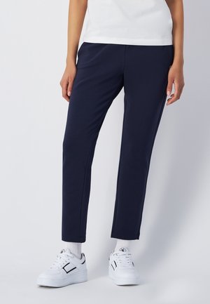 Osoba w granatowych wąskich spodniach, białych trampkach i białej bluzce, stojąca na tle jednolitego jasnego tła.