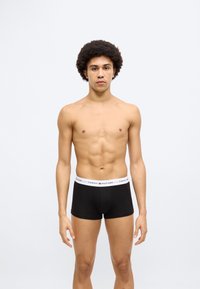 Μαύρα μποξεράκια με λευκή μέση με λογότυπο. Το μοντέλο στέκεται όρθιο μπροστά σε απλό φόντο, παρουσιάζοντας το σχέδιο και τη εφαρμογή.