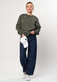 Femeie purtând un hanorac verde măsliniu, blugi largi întunecați, adidași albi, ținând o șapcă albă, stând în fața unui fundal simplu, deschis la culoare.