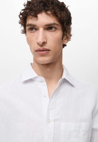 Weißes Leinenhemd mit Knöpfen, einem abgerundeten Kragen und einer Brusttasche. Verfügt über eine atmungsaktive Textur und eine entspannte Passform. Minimalistisches Design.