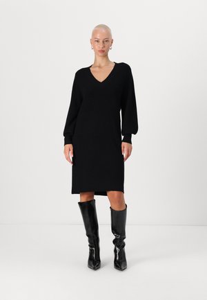 Black knitted dress with a v-neck, long puff sleeves, and straight silhouette. Accompanied by knee-high shiny black block-heeled boots.