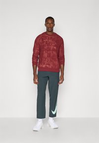 Bordový texturovaný svetr s dlouhými rukávy a kulatým výstřihem, v kombinaci s tmavozelenými sportovními kalhotami s mátovým logem Nike a bílými teniskami.