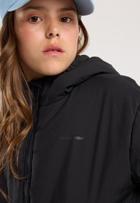 Black hooded jacket with a smooth texture, featuring a zip closure and a subtle logo on the chest. Paired with a light blue cap.