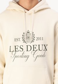 Cream-colored hoodie featuring a front pocket, with dark print of "LES DEUX Sporting Goods," est. 2011, and decorative emblem above text.