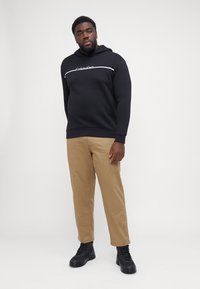 Zwarte hoodie met witte logo op de borst, gecombineerd met beige broeken en zwarte laarzen. Glad materiaal, casual pasvorm, minimalistisch ontwerp.
