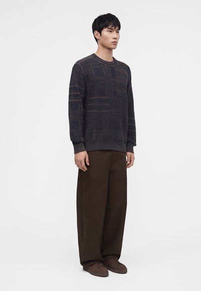 Jeune homme debout portant un pull en tricot texturé foncé, un pantalon large foncé et des baskets marron sur un fond blanc uni.