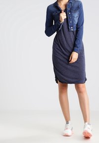 Denim cropped jacket with buttons over a dark blue jersey dress. The outfit is paired with white and pink sneakers. Smooth textures, casual style.