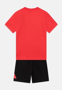 Camiseta de algodón roja con mangas cortas y cuello redondo, combinada con pantalones cortos negros que tienen un acento triangular rojo en la pierna izquierda.
