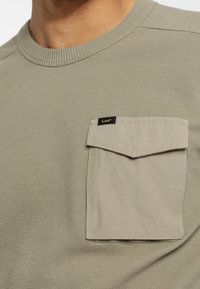 Sweat-shirt vert olive clair en tissu texturé, doté d'une poche poitrine beige avec une étiquette noire "Lee". Encolure ronde.