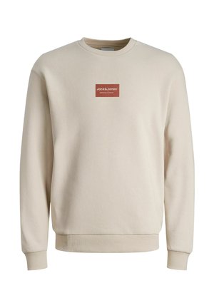 Beżowa bluza z okrągłym dekoltem, z długimi rękawami i ściągaczami przy mankietach, z małą czerwoną prostokątną naszywką z logo "Jack&Jones Definition of Denim" na piersi.