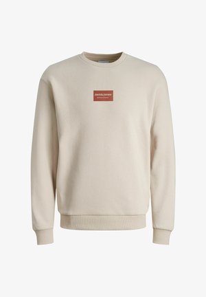 Beżowa bluza z okrągłym dekoltem, z długimi rękawami i ściągaczami przy mankietach, z małą czerwoną prostokątną naszywką z logo "Jack&Jones Definition of Denim" na piersi.