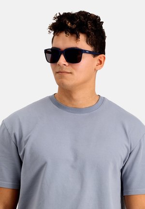 Gafas de sol con lentes oscuros y montura azul marino. Presentan una forma rectangular y un logotipo sutil. Se llevan con una camiseta gris claro.