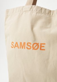 Bežs kanvas rokassoma ar uzrakstu "SAMSOE" treknajos oranžos burtos. Somai ir teksturēta virsma un izturīgas plecu siksnas.
