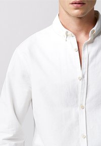 Camicia bianca con colletto aperto, realizzata in cotone. Presenta una texture liscia e una patta frontale con bottoni di colore chiaro.