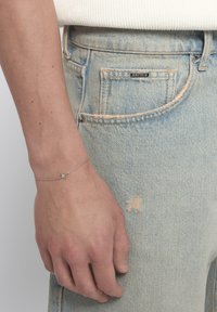 Pulsera de estrellas plateadas en una mano, llevada con unos vaqueros de denim azul claro desgastados que presentan un bolsillo visible y detalles deshilachados.