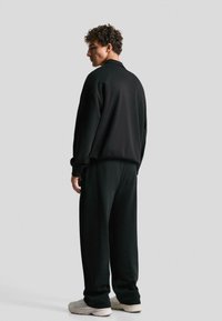 Black tracksuit made of soft fabric, featuring a relaxed fit. The model stands with back turned, showcasing the smooth texture and simple design.
