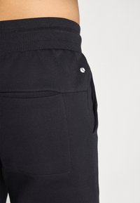 Čierne teplákové nohavice s hladkou textúrou, výrazným pásom a bočným vreckom. Malý biely logotyp na zadnej strane.