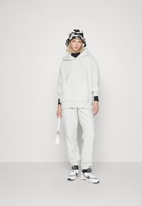 Vit fleece-hoodie med framficka och snörning, kombinerad med matchande sweatpants. En svart bucket hat med kospotsmönster och sneakers.