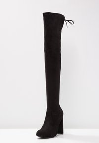 Schwarze Overknee-Stiefel aus Wildleder mit einem markanten Absatz. Mit einer Schleifen-Detail am oberen Ende und einer glatten Textur.