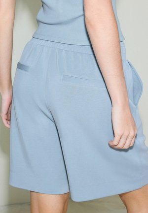 Inimene, kes kannab helesinist soonikutega pusa ja sobivaid lahtisi lühikesi pükse elastse vöökohaga ning tagaküljel asuvate pasplitega taskutega, seisab hele tausta ees.