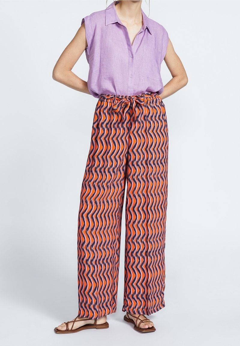 Lila ärmlös skjorta med krage, parat med vida, mönstrade byxor i orange med vågiga marinblå linjer. Inkluderar bruna sandaler med remmar.