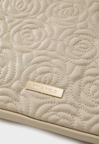 Sac à main en faux cuir matelassé beige avec un motif floral et une plaque de marque en métal doré. Texture lisse avec des détails de couture subtils.