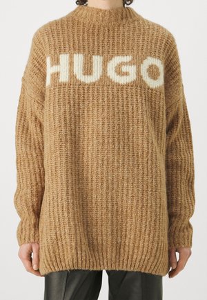 Oversizowy beżowy gruby sweter z białym napisem „HUGO” na klatce piersiowej, noszony z czarnymi skórzanymi spodniami.