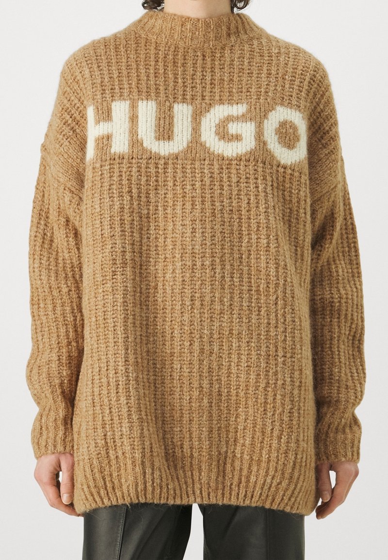 Pull en maille épaisse beige oversize avec inscription blanche "HUGO" sur la poitrine, porté avec un pantalon en cuir noir.