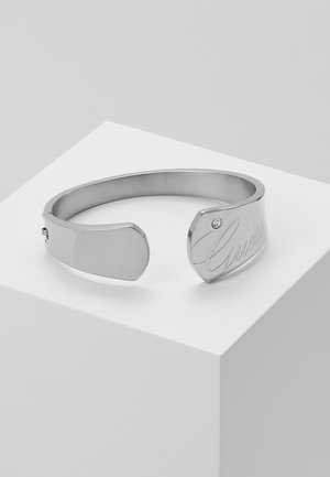 Srebrna otwarta bransoletka-mankiet z małym kryształem i wygrawerowanym napisem "Guess" na jednym końcu, prezentowana na białym piedestale.