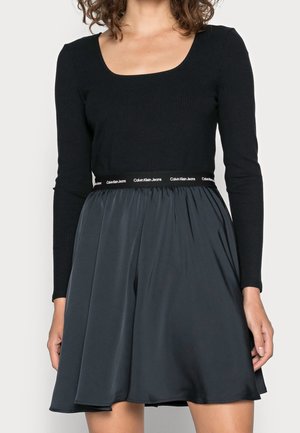 Woman wearing a black long-sleeve fitted top and a dark gray flared skirt with a Calvin Klein Jeans branded waistband.