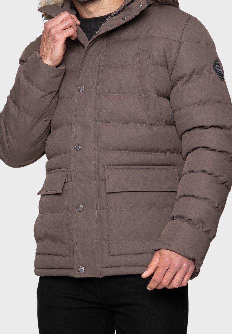 Giacca a vento marrone con cappuccio, tasche laterali e bottoni a scatto. Presenta una texture trapuntata e una piccola etichetta con logo sul petto.