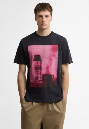 Jonge man met kort haar die een zwart t-shirt draagt met een roze geruit thermosfles en stoomkom erop, gecombineerd met beige broek.