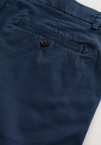 Poche arrière d'un pantalon bleu marine avec une seule fermeture à bouton noir et des coutures visibles sur un tissu lisse.
