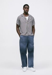 Camisa de manga curta xadrez em castanho claro, branco e azul, com corte descontraído, combinada com calças azuis largas e ténis brancos.