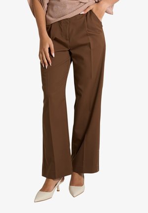Donna con pantaloni marroni a gamba dritta e décolleté beige con punta affilata, una mano in tasca e l’altra che riposa di lato.