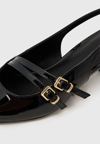 Chaussure plate en cuir verni noir avec deux brides à boucle dorée sur le devant et un design arrière ouvert avec bride.