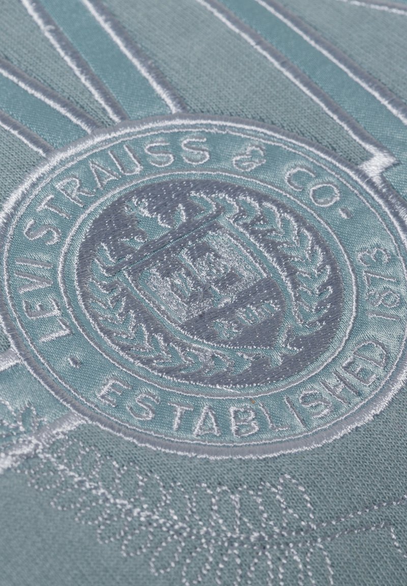 Patch ricamato rotondo con "Levi Strauss & Co." in filo argentato su uno sfondo di tessuto teal, con foglie di alloro decorative e raggi.