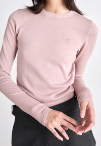 Jasnoróżowy sweter z długim rękawem wykonany z dzianiny, z okrągłym dekoltem i subtelnym logo. Posiada dopasowane rękawy oraz miękką teksturę.