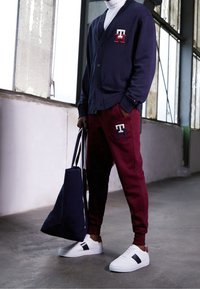 Cardigan blu navy con logo, pantaloni della tuta color amaranto con logo, sneakers bianche con dettagli neri e marroni, e una borsa a mano blu.