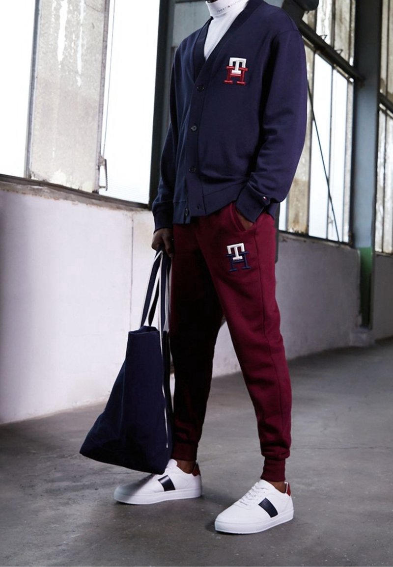 Cardigan blu navy con logo, pantaloni della tuta color amaranto con logo, sneakers bianche con dettagli neri e marroni, e una borsa a mano blu.