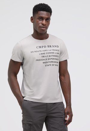 Mann trägt hellgraues T-Shirt mit schwarzem Text in Französisch und Englisch, kombiniert mit dunkelgrauen Cargohosen, steht vor einem einfarbigen Hintergrund.