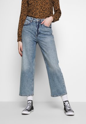 Jeans larghi azzurri in denim con una finitura scolorita, abbinati a sneakers nere alte e una camicia marrone a fantasia.