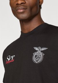 Camiseta de algodão preta com gola redonda, estampado em branco com uma águia e emblema, e texto vermelho "Ser" no lado esquerdo.