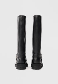 Botas de cuero negro hasta la rodilla con suelas gruesas y correas de hebilla laterales, vistas desde atrás sobre un fondo blanco liso.