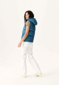 Chaleco de plumas azul con capucha, con bolsillos laterales con cremallera, sobre una camiseta de manga corta color oliva. Combinado con pantalones blancos y calzado de color claro.